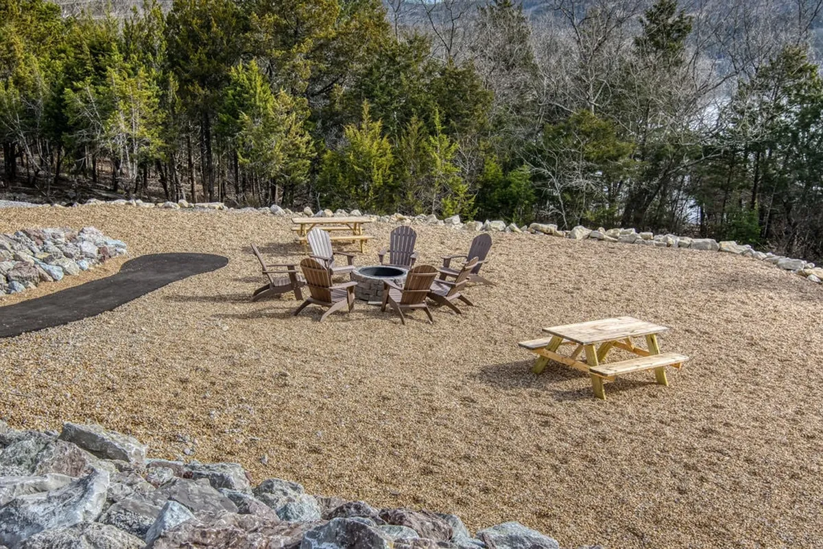 An inviting communal fire pit area surrounded by Adirondack chairs, a picnic table, and a natural pathway, set outdoors with views of the surrounding greenery. The setup encourages gatherings, storytelling, and relaxation under the open sky.