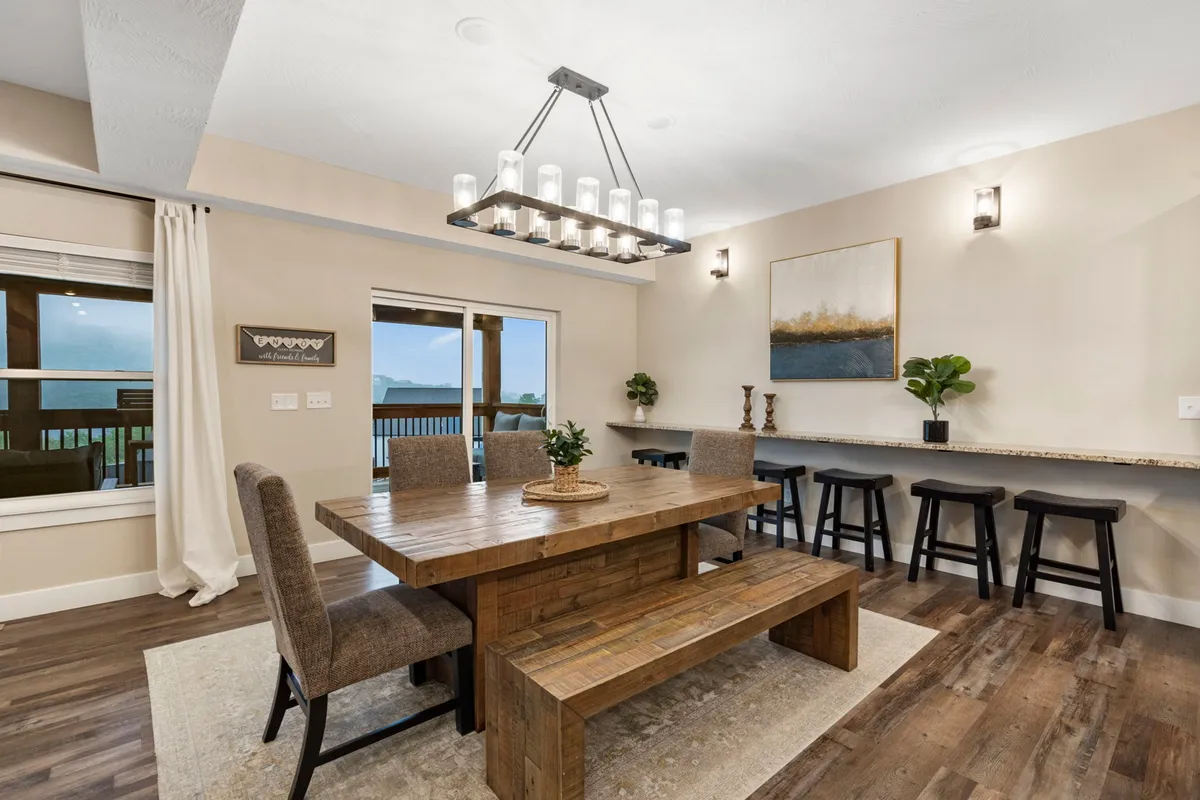A large wooden dining table set under a chandelier, with bar seating and windows offering scenic views. The area feels warm and welcoming, ideal for lively conversations and memorable meals.