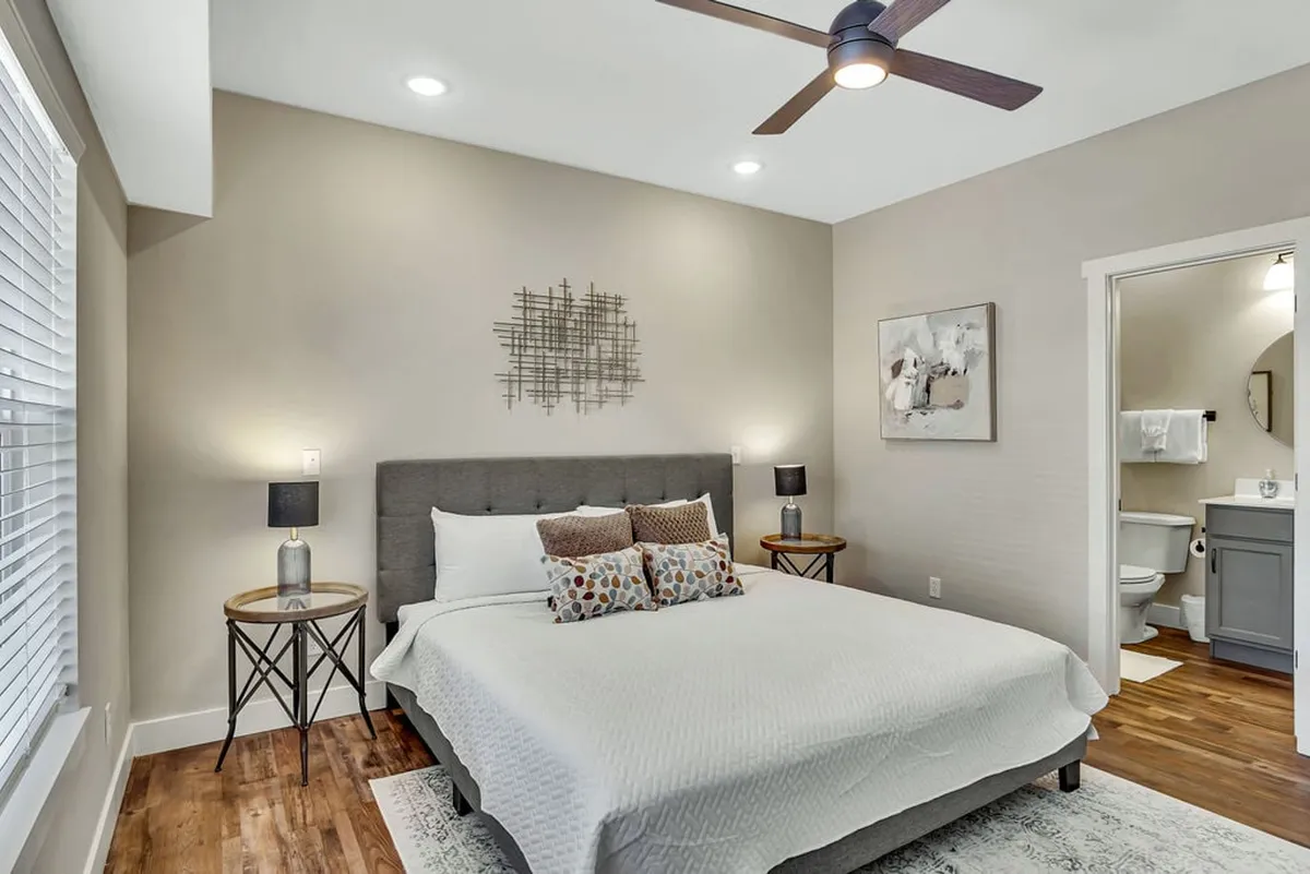 A serene and cozy king or queen suite, with a plush bed adorned with decorative pillows, nightstands with soft lamp lighting, and artwork on the wall. The suite includes a bathroom en suite visible through an open doorway, offering a peaceful atmosphere for restful nights and easy access to spa-inspired amenities.