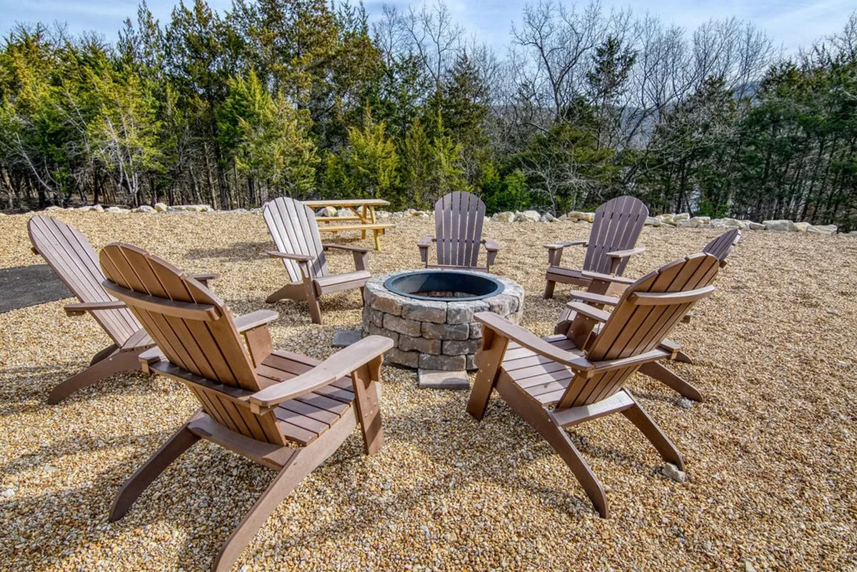 A cozy fire pit area surrounded by Adirondack chairs, set in a natural outdoor environment with trees and a gravel pathway—suggesting warmth, relaxation, and communal gatherings under the stars.