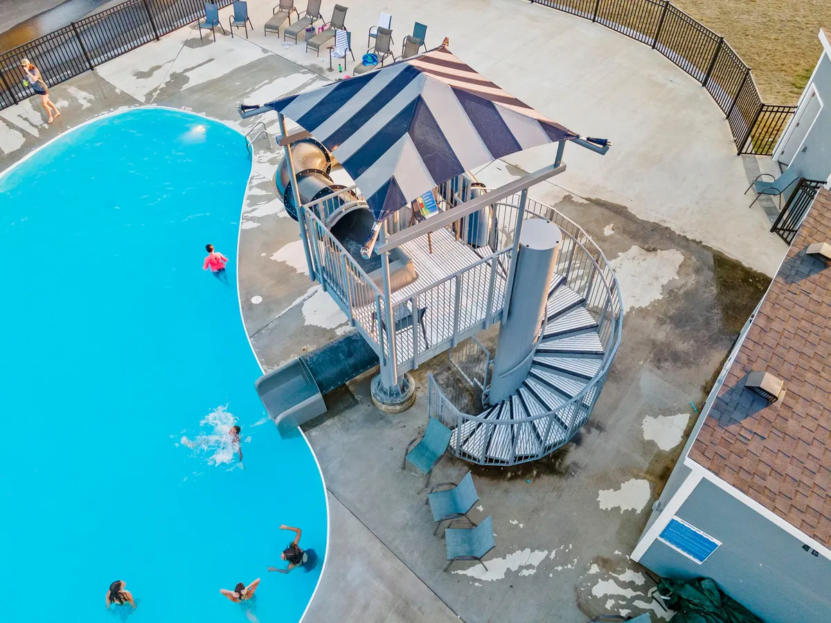 A vibrant outdoor swimming pool area featuring a water slide, poolside seating, and children playing, surrounded by greenery and sunshine. The scene captures the energy and joy of summertime family activities.