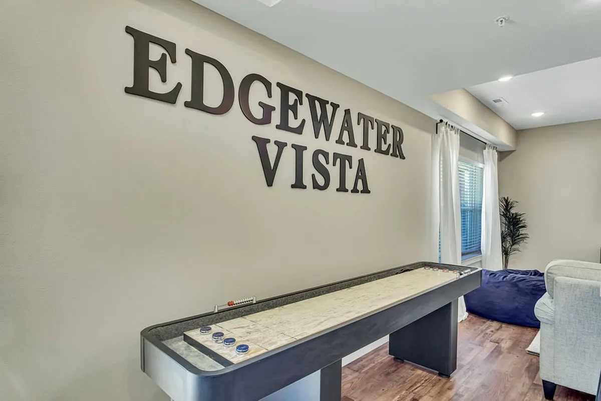 A lively interior space with a large 'EDGEWATER VISTA' wall sign, a shuffleboard table, and a comfortable seating area. The setup invites casual game nights, friendly competition, and memorable social gatherings with family and friends.