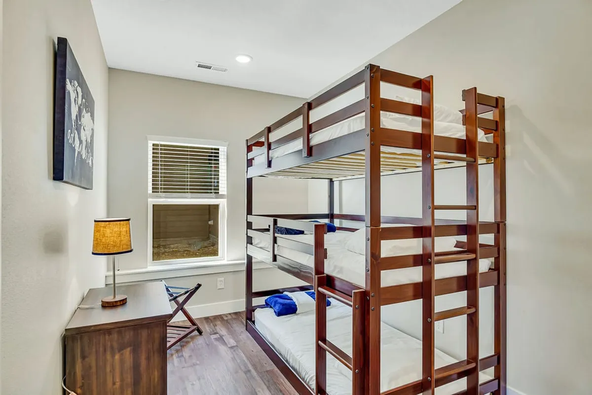 A cheerful and playful bunk room featuring a triple bunk bed, a nightstand with a lamp, and a window letting in daylight. The setup is ideal for kids or teens, creating a fun and communal sleeping space that fosters shared adventures and bedtime stories.